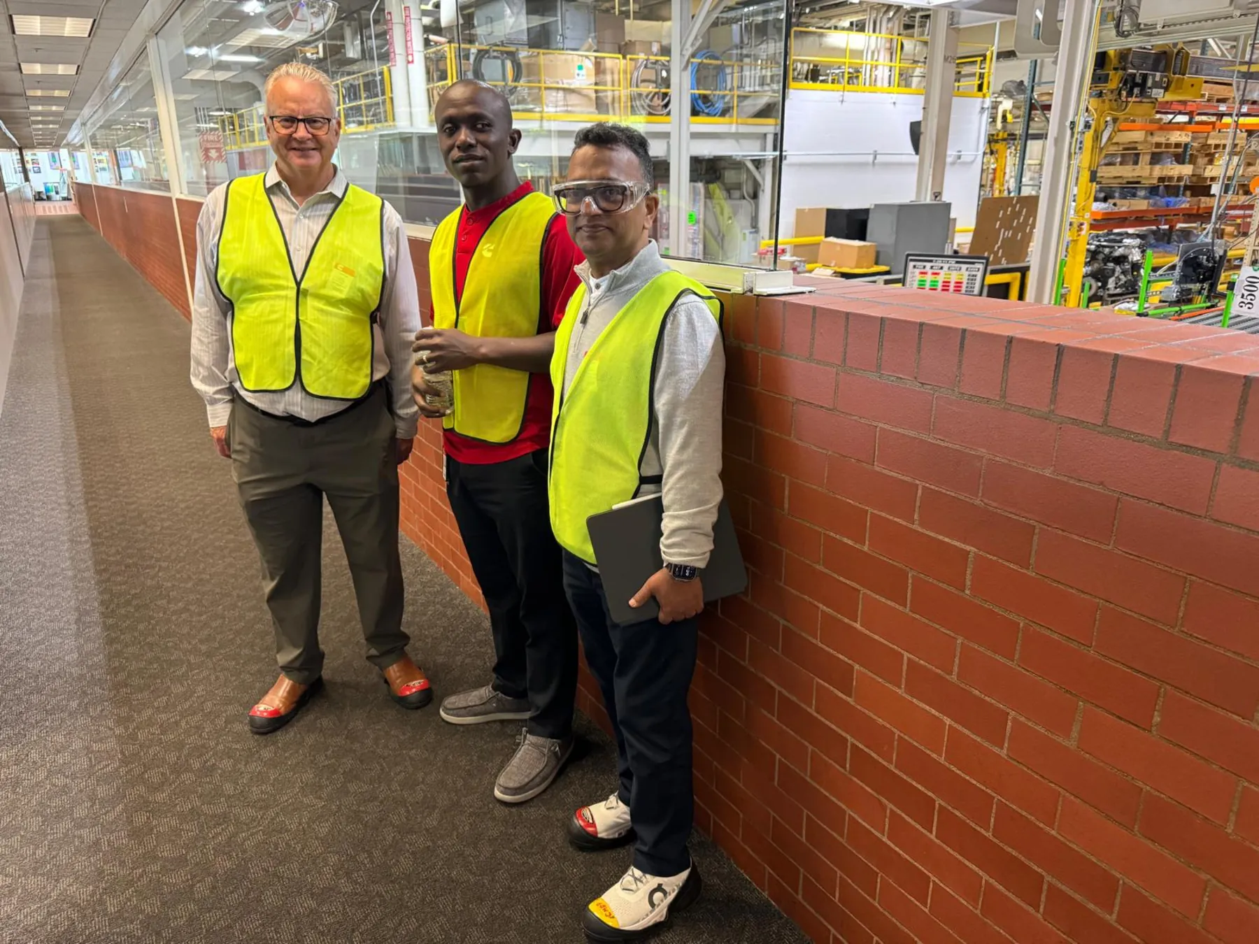 Three people in high-visibility vests on a mezzanine walkway above a working plant floor