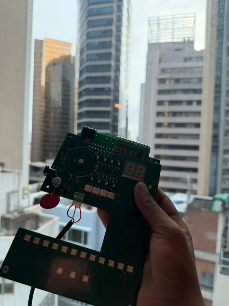A hand holding up a prototype circuit board against a window over city towers